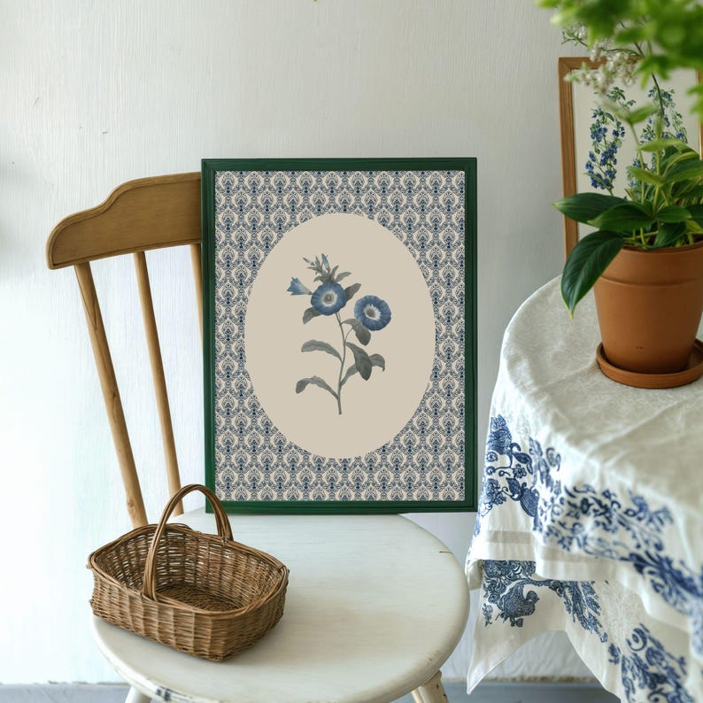 May include: A framed botanical print featuring a blue flower illustration. The artwork is set against a cream-colored oval, surrounded by a blue and white patterned background, and enclosed in a green frame. A small wicker basket sits on a white chair.
