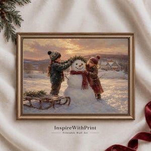 May include: A framed wall art print depicting two children in winter clothing building a snowman in a snowy landscape at sunset. The snowman wears a red scarf and a green wreath. A wooden sled sits on the snow nearby. The text InspireWithPrint appears below.