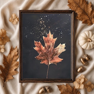 May include: A framed wall art print featuring a single copper-toned maple leaf centered against a dark charcoal background with scattered gold-colored speckles. The frame is dark brown with a textured, beaded edge detail.