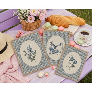 May include: Three framed botanical prints with floral and hummingbird illustrations. The prints have a beige oval center with blue floral designs and a patterned border. A picnic setting includes a basket of flowers, pastries, and a cup of coffee.