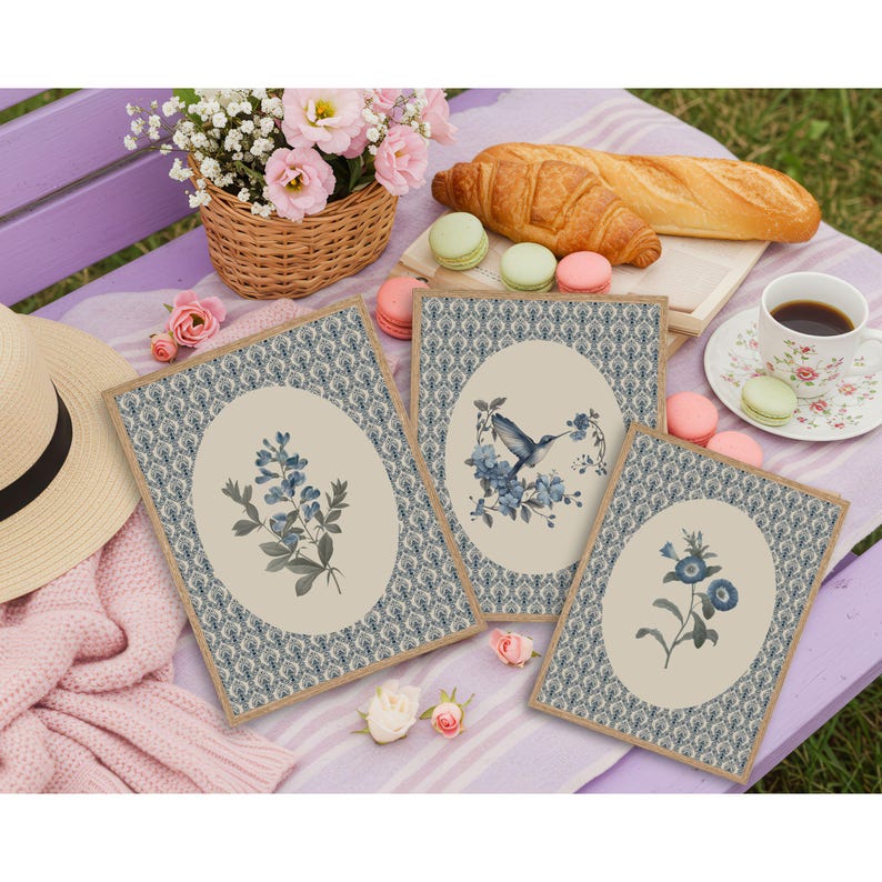 May include: Three framed botanical prints with floral and hummingbird illustrations. The prints have a beige oval center with blue floral designs and a patterned border. A picnic setting includes a basket of flowers, pastries, and a cup of coffee.
