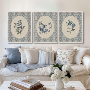May include: Set of three framed wall art prints. Each print features a botanical illustration in shades of blue and gray, set against a cream-colored oval. The prints are surrounded by a patterned border. The prints are displayed above a white sofa with blue and white pillows.