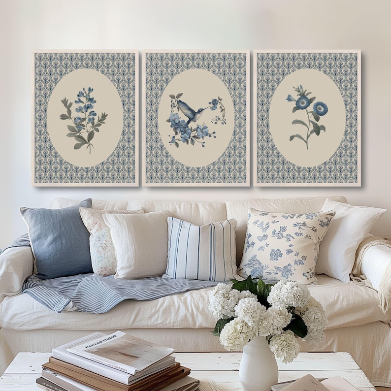 May include: Set of three framed wall art prints. Each print features a botanical illustration in shades of blue and gray, set against a cream-colored oval. The prints are surrounded by a patterned border. The prints are displayed above a white sofa with blue and white pillows.