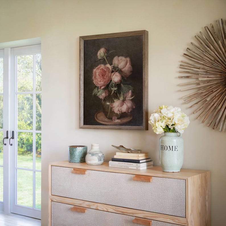 K&ouml;nnte beinhalten: Ein gerahmtes Gem&auml;lde eines Strau&szlig;es rosa Pfingstrosen in einer Vase auf einem Holztisch mit einer Vase mit wei&szlig;en Blumen, einem Stapel B&uuml;cher und zwei dekorativen Keramikbeh&auml;ltern. Das Gem&auml;lde hat einen dunklen Hintergrund und einen Holzrahmen. Der Tisch ist aus hellem Holz mit zwei Schubladen. Die Wand ist hellbeige.