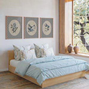May include: A light wood bed frame with a light blue duvet and pillows. Three framed botanical prints hang above the bed. The room has a large window with a view of trees and two brown vases on the windowsill.