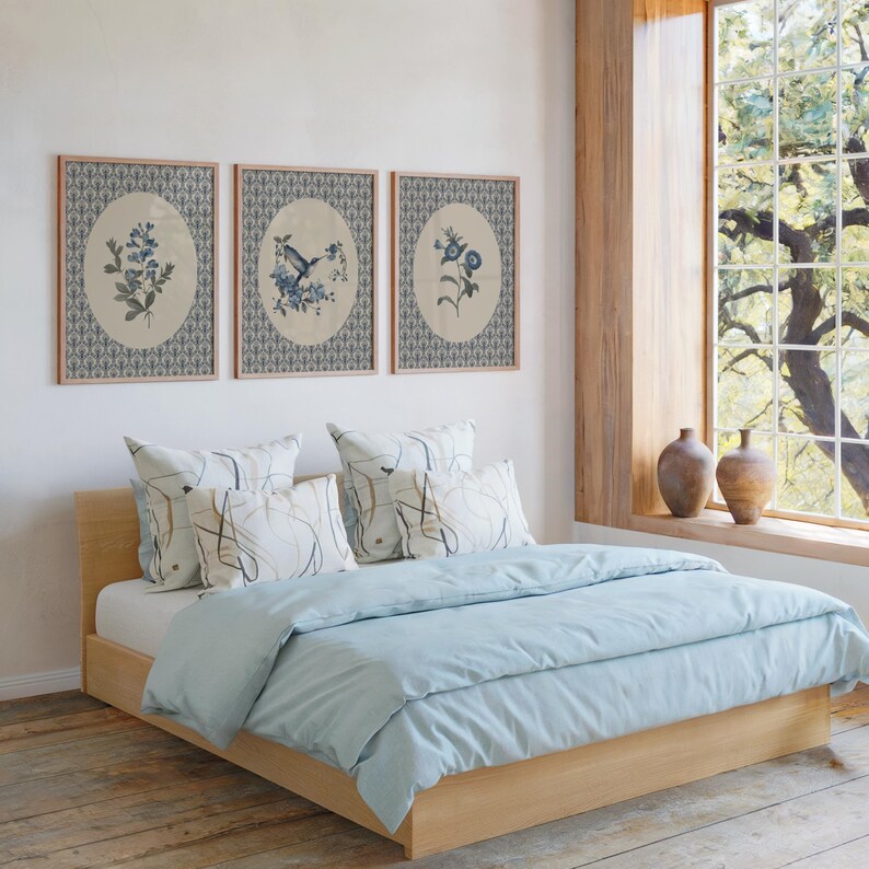 May include: A light wood bed frame with a light blue duvet and pillows. Three framed botanical prints hang above the bed. The room has a large window with a view of trees and two brown vases on the windowsill.