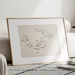 以下が含まれることがあります： ニュートラルな色合いの紙に黒インクで描かれた、飛んでいる鳥の群れを描いた額装されたアートワーク。アートワークは白いマットと明るい茶色の木製フレームに収められています。額装されたアートは、模様のあるラグの上の白いソファに寄りかかっています。
