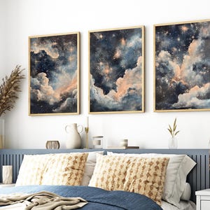 Pode incluir: Três impressões de arte emolduradas retratando uma cena celestial com nuvens rodopiantes em tons de azul, pêssego e branco, acentuadas por estrelas douradas. A obra de arte é exibida acima de uma cama com travesseiros estampados e uma cabeceira azul.