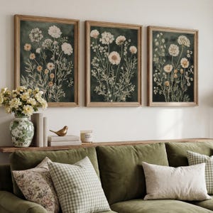 May include: Three framed botanical prints with a dark green background and light-colored floral illustrations. The frames are a light brown color. The prints are displayed above a green sofa with decorative pillows and a wooden shelf with a vase and books.