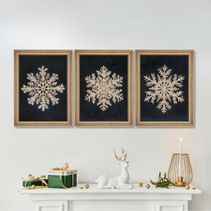 May include: Three framed prints of gold snowflake illustrations on a dark blue background. The frames are gold and the snowflakes are detailed and intricate.