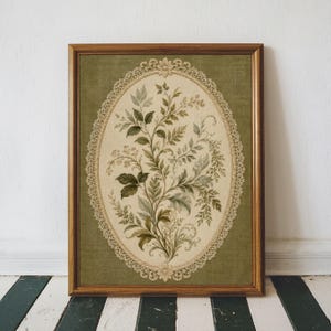 May include: A framed botanical print featuring an oval floral design. The artwork showcases a detailed arrangement of green and beige leaves and flowers, set against a green background. The print is enclosed in a wooden frame, adding a touch of vintage charm.