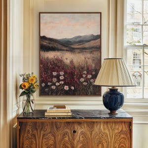 Pode incluir: Uma pintura emoldurada de uma paisagem com um campo de flores brancas em primeiro plano e uma cordilheira ao fundo. A pintura est&aacute; pendurada em uma parede acima de um arm&aacute;rio de madeira com uma l&acirc;mpada em cima.