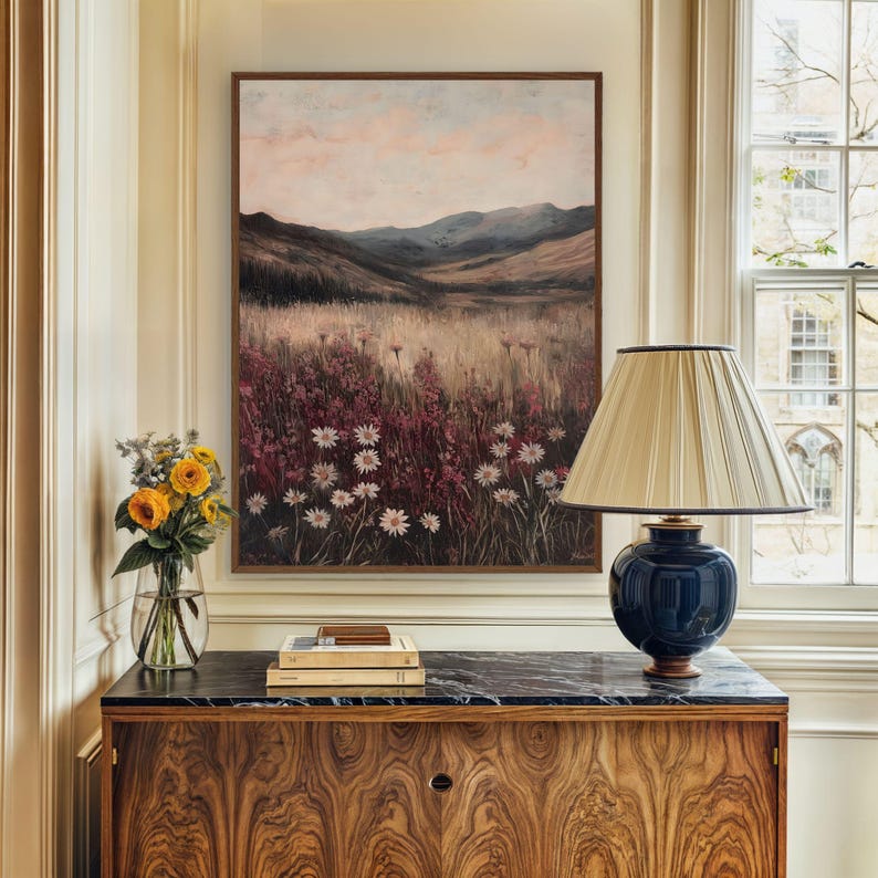 Pode incluir: Uma pintura emoldurada de uma paisagem com um campo de flores brancas em primeiro plano e uma cordilheira ao fundo. A pintura est&aacute; pendurada em uma parede acima de um arm&aacute;rio de madeira com uma l&acirc;mpada em cima.