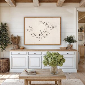 以下が含まれることがあります： リビングルームのシーン。鳥の群れが飛んでいる大きな額入りのアートワーク。アートワークは、木製のアクセントが付いた白いキャビネットの上に掛けられています。木製のコーヒーテーブルと鉢植えの植物が装飾を完成させています。