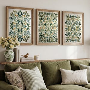 Può includere: Tre stampe artistiche botaniche incorniciate con un design simmetrico di foglie nei toni del verde, blu e giallo. Le stampe sono esposte sopra un divano verde con cuscini decorativi, un vaso di fiori e una piccola statuetta di uccello dorato.