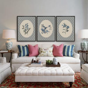 May include: Three framed floral prints with black frames hang above a white sofa with pink and blue pillows. The prints feature blue floral designs on an oval cream background with a patterned border. A white ottoman and patterned rug complete the room.