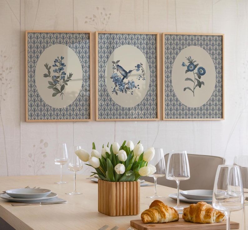 May include: Three framed botanical prints with oval center images of blue flowers and a hummingbird. The frames are light wood, and the background is a blue and white patterned design. A dining table is set with white tulips, plates, and glasses.