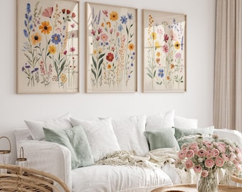 May include: Three framed botanical prints featuring a variety of colorful wildflowers on a cream background. The prints are hung on a white wall above a white couch with throw pillows and a blanket.