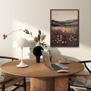 Pode incluir: Uma mesa de madeira com uma l&acirc;mpada branca, um vaso com flores, um laptop, uma x&iacute;cara e um pires, e um livro. Uma pintura emoldurada de uma paisagem com montanhas e flores silvestres pendura na parede atr&aacute;s da mesa.