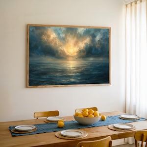 May include: A framed seascape painting with a dramatic sky. The artwork shows a sunset over the ocean, with blue, gold, and grey hues. The painting hangs above a wooden dining table set with plates, a blue runner, and a bowl of lemons.