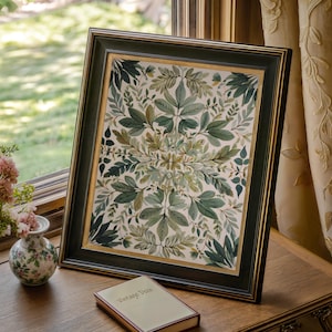 Könnte beinhalten: Ein gerahmter botanischer Druck mit einem symmetrischen Muster aus grünen Blättern. Der Rahmen ist dunkelgrün mit einem goldenen Innenrand. Eine kleine Vase mit rosa Blumen und ein Buch mit der Aufschrift "Vintage Deco" sind ebenfalls vorhanden.