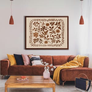 May include: A brown framed print with a cream background featuring a floral design in shades of brown. The print is hanging on a white wall above a brown sectional sofa with yellow throw pillows and a blanket.