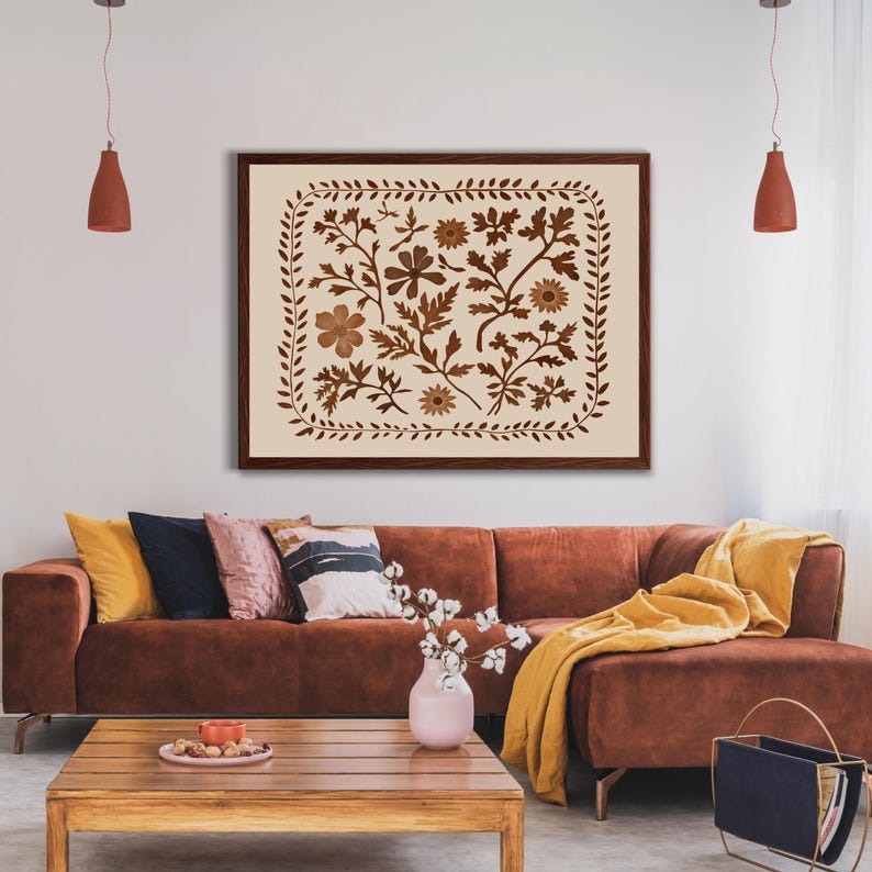 May include: A brown framed print with a cream background featuring a floral design in shades of brown. The print is hanging on a white wall above a brown sectional sofa with yellow throw pillows and a blanket.