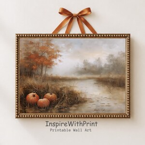 May include: A rectangular wall art print in a beaded gold frame, suspended by a copper-colored ribbon. The artwork depicts a misty autumn landscape with three orange pumpkins in the foreground, tall grasses, and trees with orange foliage by a pond.