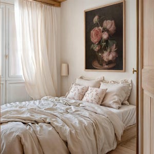 K&ouml;nnte beinhalten: Ein beigefarbener Leinen-Bettbezug mit einem wei&szlig;en, durchsichtigen Vorhang im Hintergrund. Das Bett ist mit zwei beigen Leinenkissen und zwei Kissen mit Blumenmuster bezogen. Ein gerahmtes Gem&auml;lde mit rosa Rosen in einer Vase h&auml;ngt an der Wand &uuml;ber dem Bett.