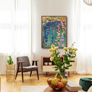 May include: A framed abstract painting of a colorful floral garden with pink, yellow, and blue flowers. The painting is hanging on a white wall in a living room setting with a gray armchair, a wooden side table, and a vase of white flowers.