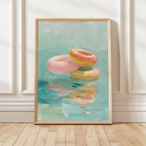 May include: A framed art print depicting three inflatable rings in water. The rings are pink, orange, and yellow, with reflections in the water. The background is a light blue, creating a tranquil summer scene. The frame is made of wood.