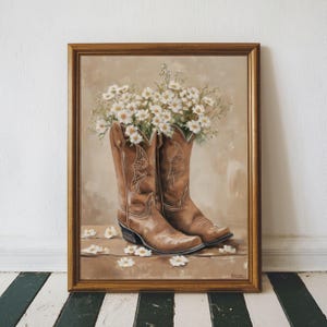 May include: A framed painting shows brown cowboy boots filled with white daisies. The boots feature decorative stitching. White daisies are scattered around the base. The painting has a warm, rustic aesthetic, with a brown and beige colour palette.