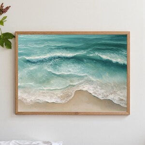 May include: A framed art print depicting an aerial view of ocean waves crashing onto a sandy beach. The artwork features shades of turquoise and white, with a light brown wooden frame. The scene evokes a sense of tranquility and the beauty of the coast.