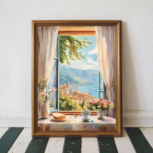 Op de afbeelding: Een ingelijst schilderij toont een schilderachtig uitzicht door een raam. Het kunstwerk toont een kustplaats met bergen, een blauwe zee en een heldere lucht. Een tafel voor het raam heeft een kopje thee, bloemen en brood.