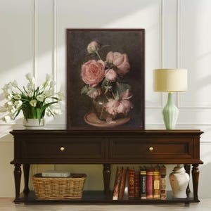 K&ouml;nnte beinhalten: Ein gerahmter Vintage-Stillleben-Druck mit einem Strau&szlig; rosa Pfingstrosen in einer Glasvase auf einem Holztisch. Der Druck ist vor einem dunklen Hintergrund platziert und hat eine rustikale &Auml;sthetik.