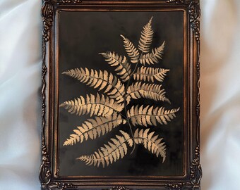 May include: A framed botanical print featuring a gold fern frond on a black background. The fern is centered within a dark bronze-colored ornate frame. The text "InspireWithPrint Printable Wall Art" is at the bottom.