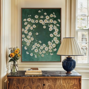 May include: A framed print of a wreath of white daisies on a dark green background. The print is hanging on a wall above a wooden dresser with a black marble top. A blue lamp with a gold lampshade is on the dresser.