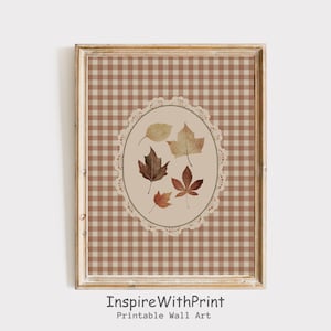 May include: A framed wall art print with a brown and beige gingham pattern background. An oval, lace-edged design in the centre displays watercolour illustrations of autumn leaves in brown and beige. The text "InspireWithPrint Printable Wall Art" is at the bottom.