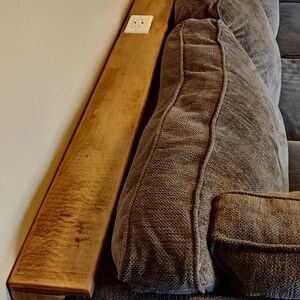 May include: A wooden shelf attached to the back of a grey sofa. The shelf has a white electrical socket on the wall behind it.