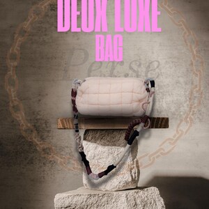 May include: A light pink quilted bag with a multi-colored strap is displayed on a stone pedestal. The bag is rectangular with a quilted pattern. The strap features white, burgundy, and black sections. Text on the image reads "The New Deux Luxe Bag".