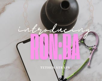 Könnte beinhalten: Ein Bild mit einem schwarzen Telefon, einem schwarzen Haltegurt und einem dekorativen schwarzen Objekt. Der Text "introducing RON:BA TETHER STRAPS" wird in Pink und Weiß angezeigt.