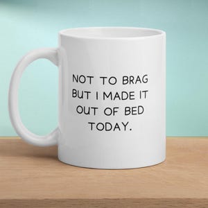 Könnte beinhalten: Weißer Keramikbecher mit schwarzem Henkel. Auf dem Becher steht der Text "NOT TO BRAG BUT I MADE IT OUT OF BED TODAY." Der Becher steht auf einer Holzoberfläche mit hellblauem Hintergrund.