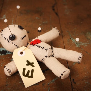 May include: A white cloth voodoo doll with black stitching and a red heart on its chest is pinned with white pins to a wooden surface. A yellow tag with the letter "E" written on it is attached to the doll.