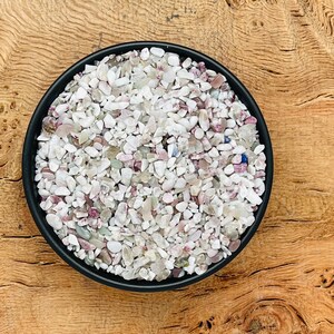 May include: A black bowl filled with small, white, pink, and blue tumbled stones. The stones are arranged in a random pattern and are all about the same size.