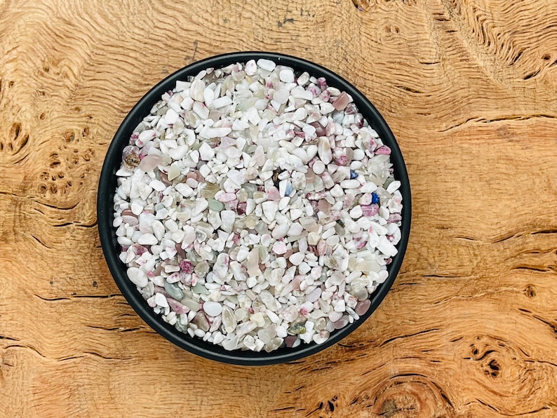 May include: A black bowl filled with small, white, pink, and blue tumbled stones. The stones are arranged in a random pattern and are all about the same size.