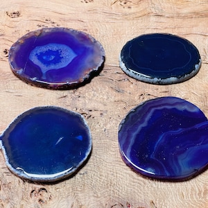 Puede incluir: Cuatro rodajas de ágata pulidas en tonos azules y morados, con patrones de bandas únicos. Cada rodaja tiene un borde natural e irregular. Las piedras se exhiben sobre una superficie de madera, mostrando su belleza natural.