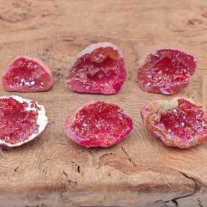 Puede incluir: Seis mitades de geoda de cristal de ágata roja iridiscente con una textura natural, cruda y rugosa. Las mitades de geoda están dispuestas sobre una superficie de madera.