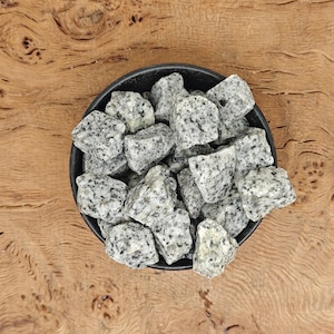 Puede incluir: Un cuenco negro lleno de rocas de granito grises y blancas. Las rocas tienen un patrón moteado y son de diferentes tamaños. El cuenco está sobre una superficie de madera con vetas naturales.