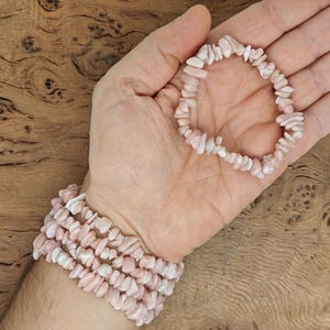 Peut inclure: Un bracelet en pierres gemmes roses et blanches, avec un aspect délicat et naturel. Le bracelet est composé de petites pierres gemmes de forme irrégulière, enfilées sur un cordon élastique transparent. Le bracelet est présenté sur le poignet d'une personne, la main ouverte et la paume vers le haut.