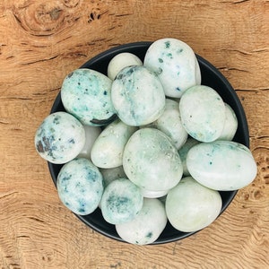 Puede incluir: Un cuenco negro lleno de piedras lisas y ovaladas. Las piedras son predominantemente blancas con áreas de color azul-verdoso y manchas oscuras. El cuenco descansa sobre una superficie de madera con un patrón de vetas visible.